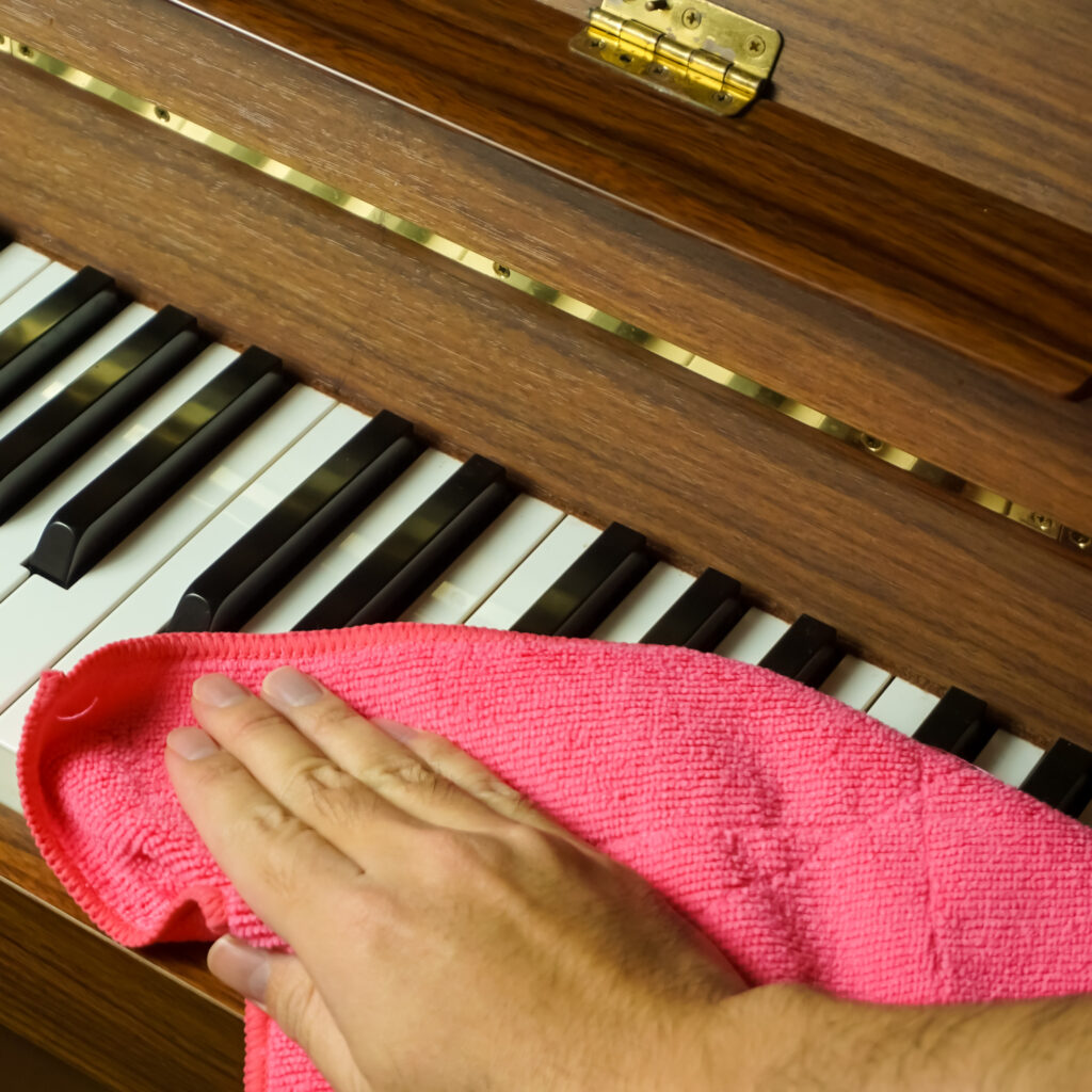 Main nettoyant les touches blanches et noires d'un piano en bois avec un chiffon microfibre rose. Détails en laiton visibles.