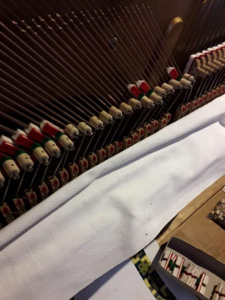 Entretien piano : mécanique interne d'un piano droit. Intérieur de piano droit avec cordes et marteaux feutrés rouges et verts. Un tissu blanc protège la mécanique, à côté d'une boîte de petites pièces.