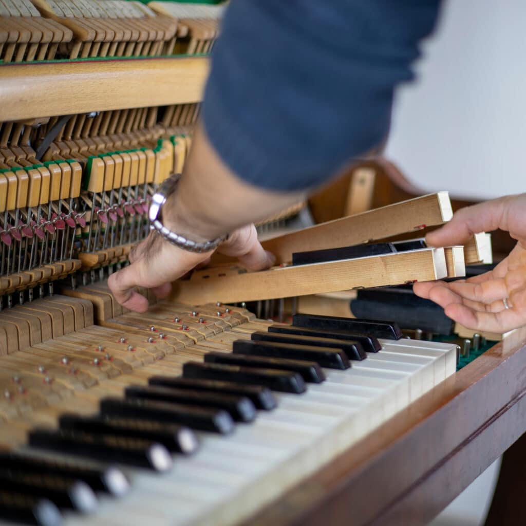 Mains ajustant le mécanisme interne d'un piano ouvert. Marteau, étouffoirs et touches visibles pendant la réparation.