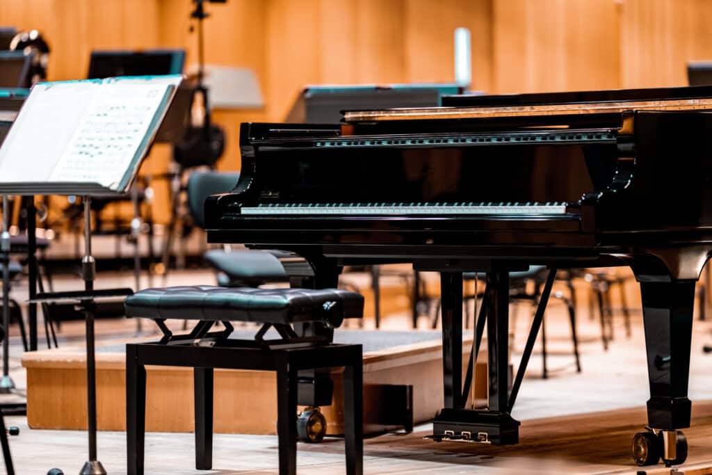Piano à queue noir brillant au premier plan, avec tabouret rembourré et partitions sur pupitre à gauche, ambiance chaleureuse.