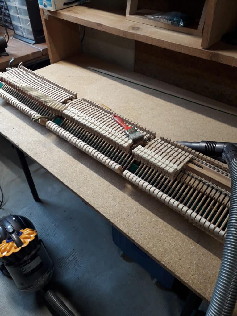 Nettoyage Mécanisme Piano : Action et Marteaux Mécanisme d'action de piano démonté, nettoyé avec un aspirateur Dyson Ball et une brosse sur un établi en aggloméré.